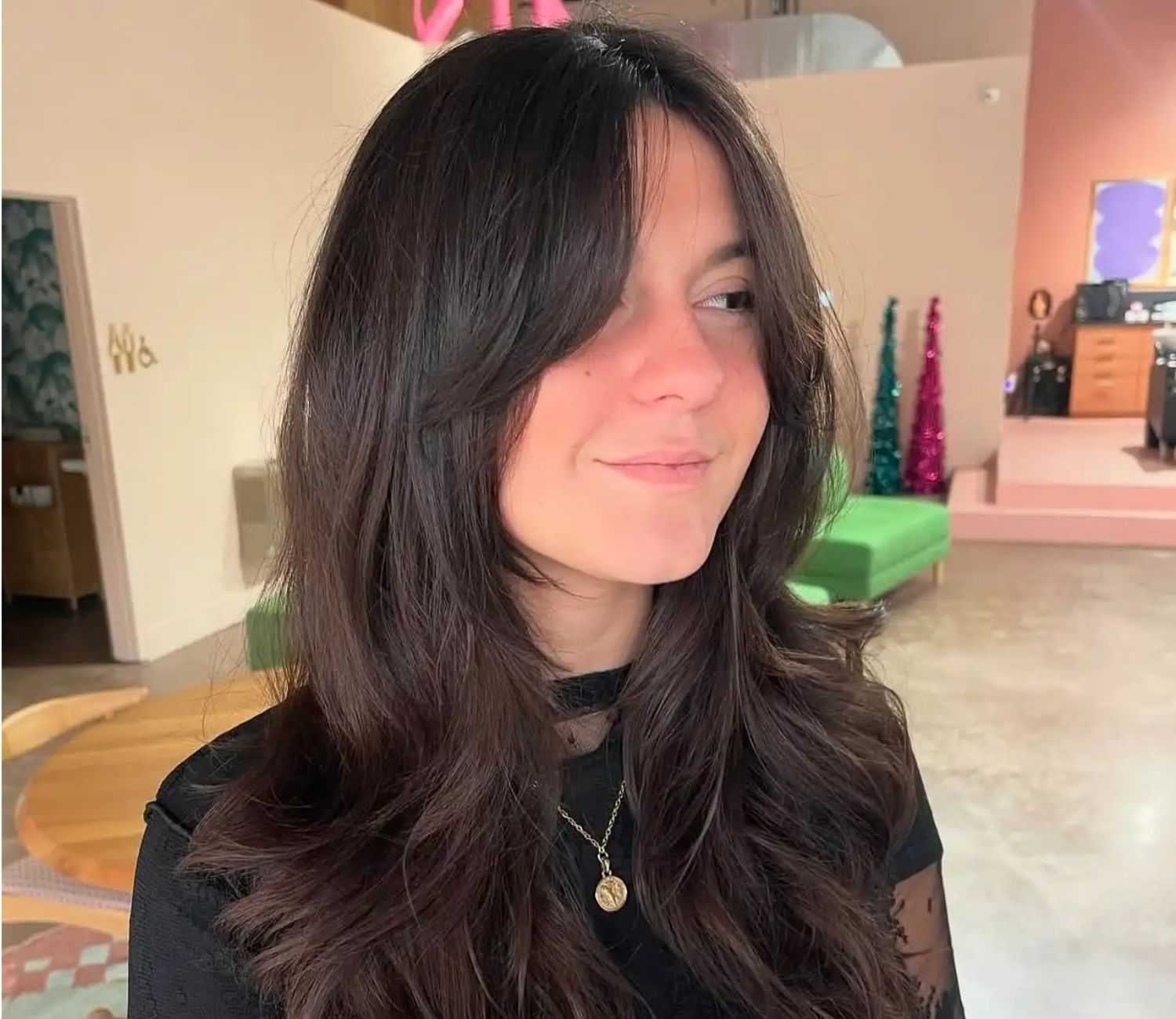 Woman with long dark hair and bangs, wearing a black top and necklace, in a modern room.