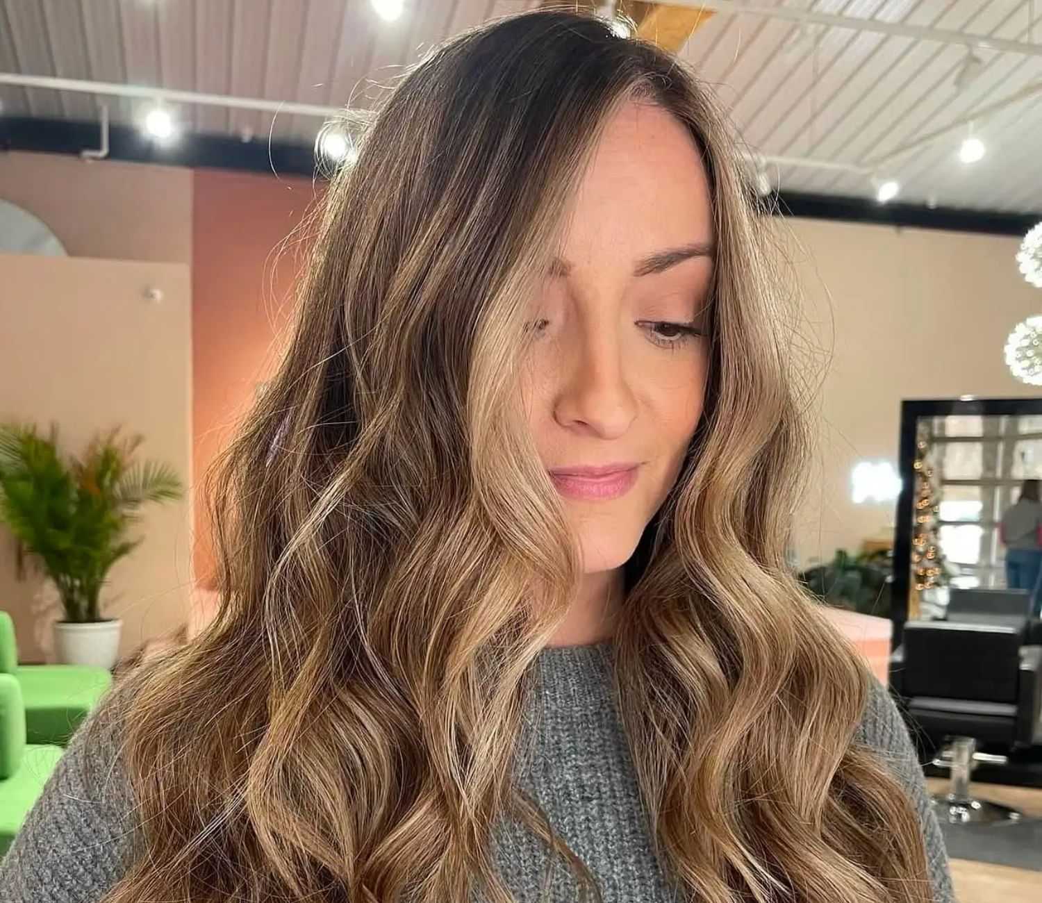 Woman with wavy, highlighted hair in a modern salon with bright lighting and plants.