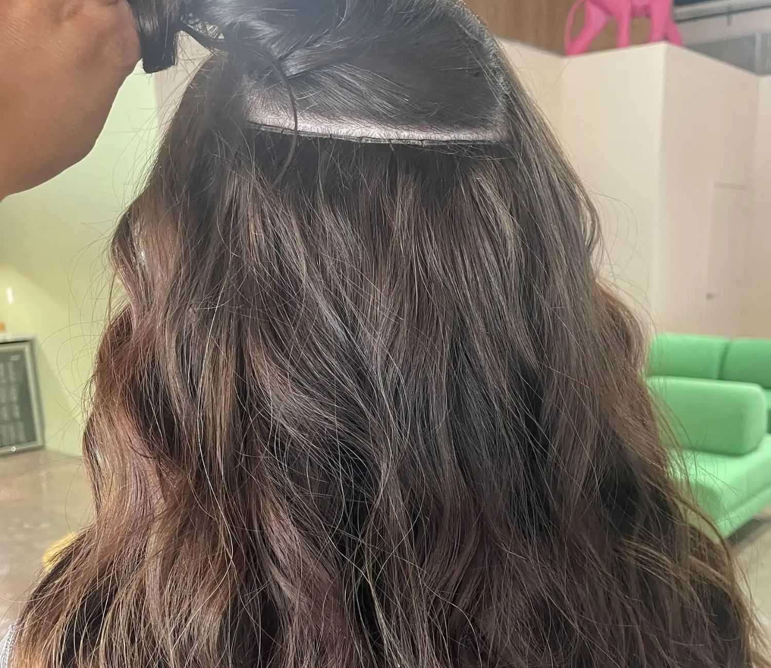 Hairstylist applying a small dark brown hair extension on a woman's wavy brown hair.